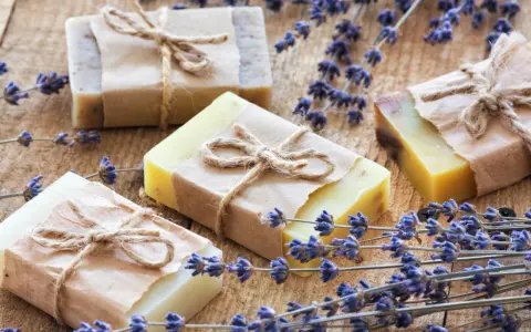 Quattro saponette fatte in casa, avvolte in carta e legate con spago, giacciono su un tavolo di legno rustico, circondate da rametti di lavanda profumata.