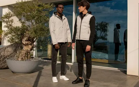 Due uomini con un moderno stile outfit uomo, uno con giacca grigia e jeans, l'altro con gilet grigio e pantaloni sportivi neri.
