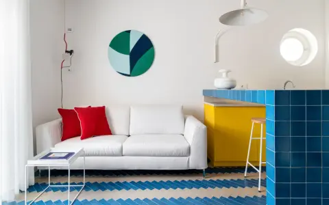 Sofá blanco con cojines rojos, mesa auxiliar blanca y suelo con diseño de espiga en colores claros y azul. Barra de cocina amarilla y azul.
