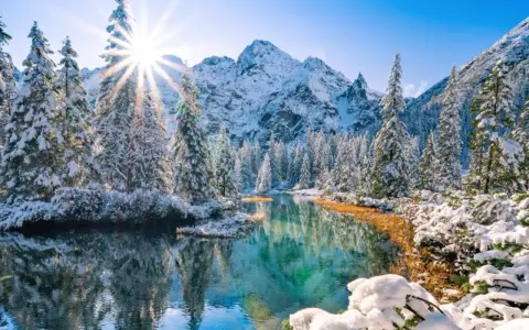 Morskie Oko zimą. Słońce przebija się przez ośnieżone drzewa, odbijając się w turkusowej tafli wody. W tle majestatyczne, pokryte śniegiem góry.