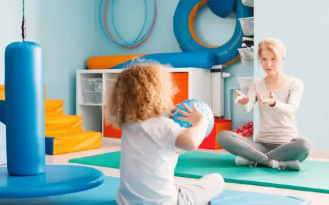 Dziecko z kręconymi włosami trzyma niebieską piłkę, siedząc naprzeciwko kobiety w pokoju terapeutycznym.