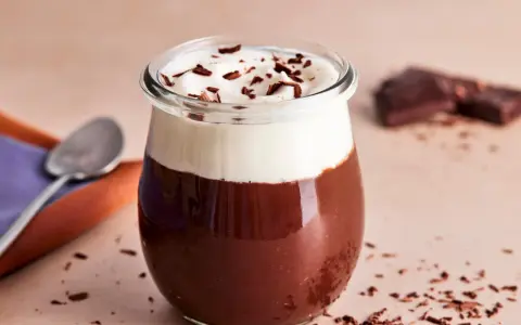 Ein Glas mit Omas Schokoladenpudding mit Sahne und Schokostreuseln. Daneben liegen ein Löffel und ein Stück Schokolade.