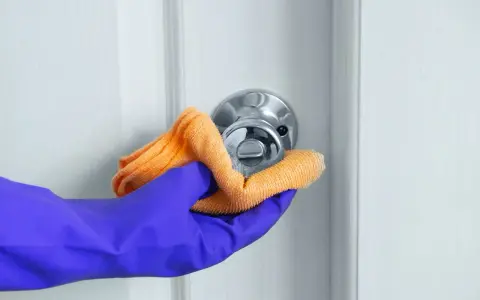 Ręka w fioletowej rękawiczce czyści klamkę pomarańczową ściereczką.