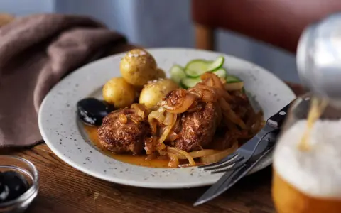 En tallrik med pannbiff, stekt lök, kokt potatis, gurka och en sked med oliver. Ett glas öl hälls upp i bakgrunden. Ett klassiskt pannbiff recept.