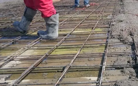 Pracownicy w gumowcach stąpają po siatce zbrojeniowej, przygotowując zbrojenie stropu do zalania betonem.