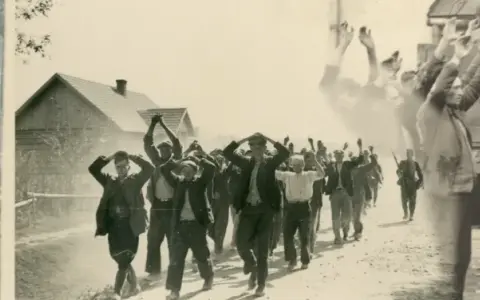 Zatrzymani mężczyźni z rękami w górze na drodze, być może w rejonie granica polsko-niemiecka 1939 Pomorze. W tle domy i żołnierz.