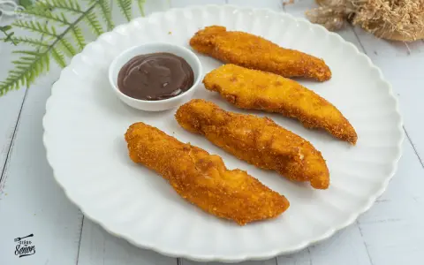 Cuatro crujientes fingers de pollo dorados, servidos en un plato blanco con una pequeña taza de salsa barbacoa.