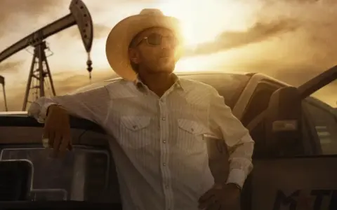 Uomo con cappello da cowboy e camicia a righe appoggiato a un pick-up, con pozzi petroliferi sullo sfondo. Il western significato è qui, tra lavoro e orizzonti infiniti.