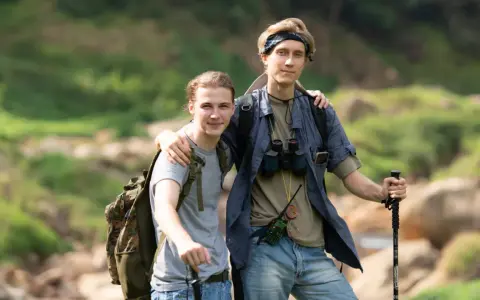 Dwóch młodych mężczyzn, gotowych na trekking, z plecakami i kijkami, w otoczeniu zieleni i skał.