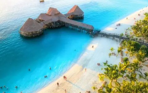 Zanzibar w czerwcu: idealna pogoda na wypoczynek na rajskiej plaży z turkusową wodą i palmami. Widać hotel na wodzie i ludzi kąpiących się.