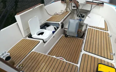 Ein weißes **Segelboot Toilette** mit offenem Deckel, daneben ein grauer Tank. Holzdecks und weiße Wände.