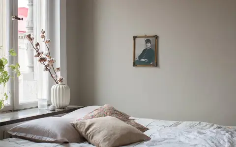 En säng med flera kuddar i olika nyanser av beige och mönster. En gammal porträttbild hänger på väggen.