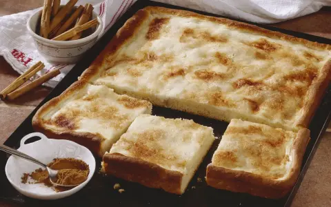 Ein köstlicher Rahmkuchen vom Blech, bestreut mit Zimt und Zucker. Daneben eine Schale mit Zimtstangen und Zimtpulver.