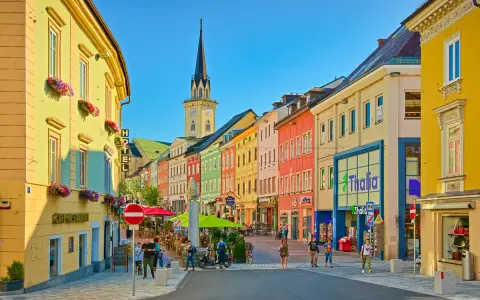 Via colorata di una città austriaca più vicina all'Italia, con edifici vivaci, una torre dell'orologio e gente che passeggia.