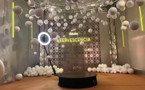 Plataforma giratoria para fotos con luces de neón "EFERVESCENCIA" y esferas blancas colgantes, creando un efecto de bullet time.