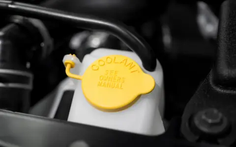 Close-up of a car's coolant reservoir with a yellow cap. The cap reads "COOLANT SEE OWNERS MANUAL," indicating where to find instructions on how to put coolant in car.