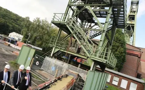 Drei Männer in Anzügen und Schutzhelmen inspizieren das Gelände der Zeche Bottrop. Ein Förderturm ragt in den Himmel.