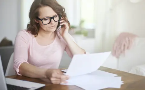 Kobieta w okularach rozmawia przez telefon, przeglądając dokumenty. Martwi się, że czeka ją brak świadectwa pracy.