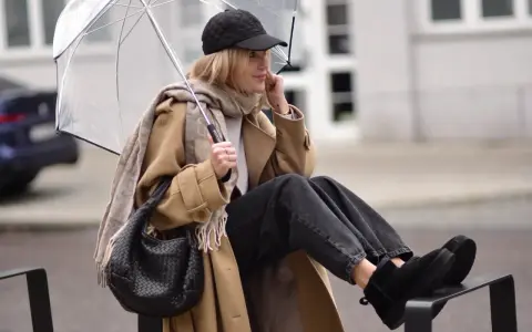 Kobieta w beżowym płaszczu, czapce i czarnych jeansach prezentuje modne emu stylizacje, trzymając przezroczysty parasol.