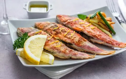 Cuatro salmonetes al horno, dorados y jugosos, servidos con rodajas de limón, perejil y patatas fritas.