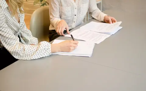 Dwie kobiety przy stole omawiają dokumenty. Jedna pisze, druga przegląda papiery. Może to umowa o zakazie konkurencji.