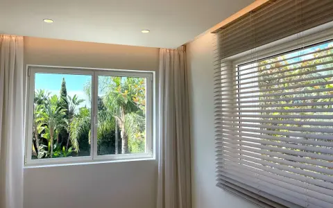 Dos tipos de apertura de ventanas: una corredera con cortinas y otra con persianas venecianas, ambas con vistas a un exuberante jardín tropical.