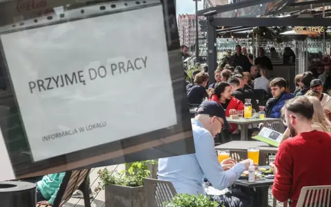 Ogłoszenie "Przyjmę do pracy" na tle ogródka restauracyjnego z ludźmi przy stolikach.