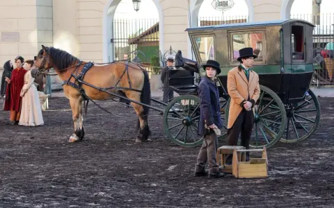 Konny zaprzęg z dorożką i postaciami w strojach z epoki na tle budynków.
