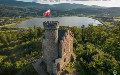 Kamienny zamek księcia Henryka z polską flagą na wieży, otoczony lasem, z widokiem na jezioro i góry. Punkt widokowy zaprasza do podziwiania krajobrazu.