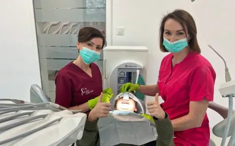 Dwie dentystki w maseczkach i rękawiczkach z uśmiechniętym pacjentem w gabinecie stomatologicznym.