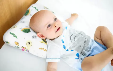 Uśmiechnięty bobas leży na białej pościeli, opierając główkę na poduszce ortopedycznej dla niemowląt z motywem żółwi.