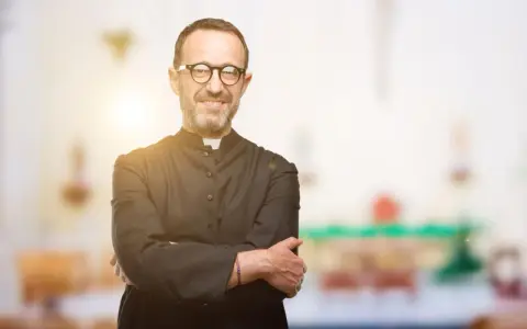 Ein lächelnder evangelischer Pfarrer mit Brille und überkreuzten Armen steht vor einem unscharfen Kircheninterieur.