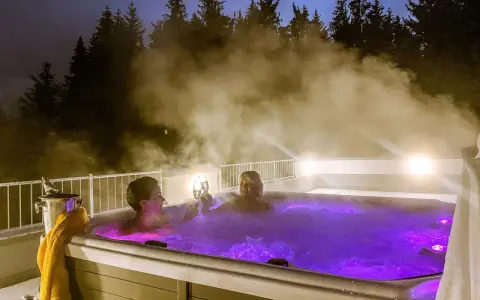 Zwei Frauen stoßen mit Champagner in einem beleuchteten Whirlpool an, umgeben von Dampf und Bäumen. Ein perfektes Wochenende mit Gott.