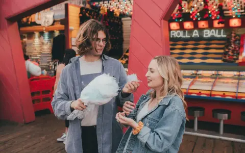 Ein Paar teilt sich Zuckerwatte vor einem "Roll a Ball"-Spiel im Freizeitpark. Ihr lässiger Freizeitpark-Outfit passt perfekt zum Tag.