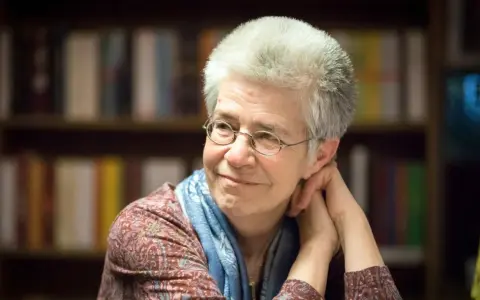 Ältere Frau mit Brille und blauem Schal lächelt nachdenklich vor Bücherregal.