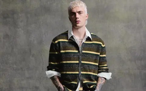Joven con pelo rubio platino y tatuajes lleva un jersey de rayas verdes y amarillas, mostrando que **que jerseys se llevan este año** son los de estilo retro.