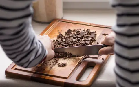 Une personne utilise un couteau pour comment casser une tablette de chocolat en pépites sur une planche à découper en bois.
