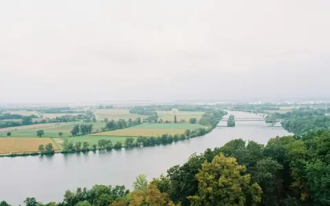 Szeroka rzeka na tle zielonych pól i lasów. W oddali widać most i zabudowania.