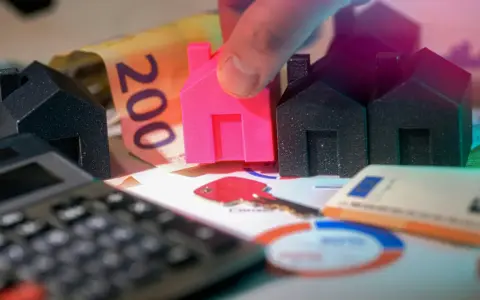 Petites maisons, billets de banque et calculatrice symbolisant le déblocage des fonds pour un projet immobilier. Une main place une maison rose.