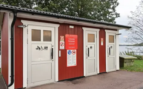 Röd byggnad med toaletter vid en sjö. Skyltar visar att camping är förbjuden. En trevlig rastplats med toalett för resenärer.