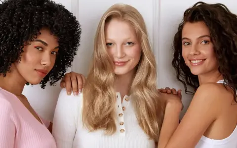 Drei Frauen stehen nebeneinander. Die Frau in der Mitte hat orange blonde haare und lächelt.
