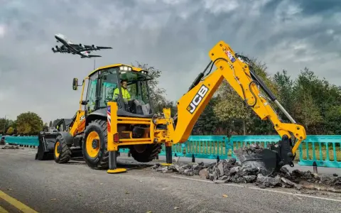 Żółta koparka JCB pracuje na drodze, a nad nią przelatuje samolot. Ciekawe, ile pali koparka, gdy pracuje tak wydajnie.