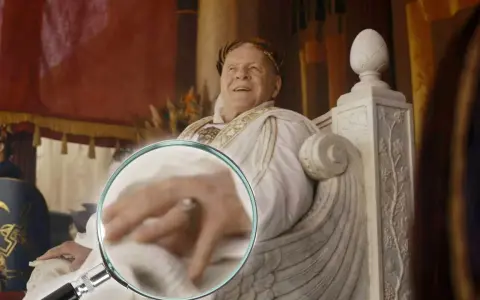 Ein Mann mit Lorbeerkranz, der an alle Kaiser Roms erinnert, sitzt auf einem Thron. Eine Lupe enthüllt Details seiner Hand.
