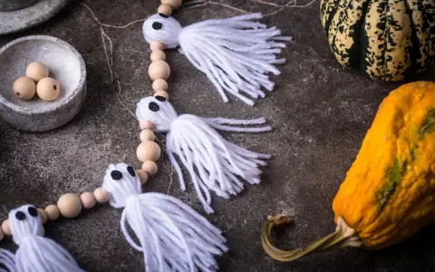 Niedliche Geister-Girlande zum Halloween basteln, mit Holzperlen und weißen Fransen. Daneben liegen bunte Kürbisse.