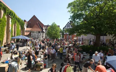 Buntes Treiben auf dem Flohmarkt Minden. Menschen stöbern an Ständen mit Kleidung und Trödel vor Fachwerkhäusern.