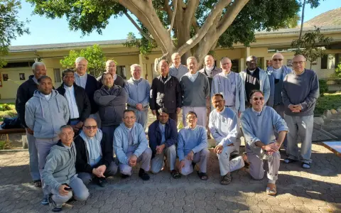 A group of missionaries of charity fathers pose for a photo outdoors, standing and kneeling under a large tree.
