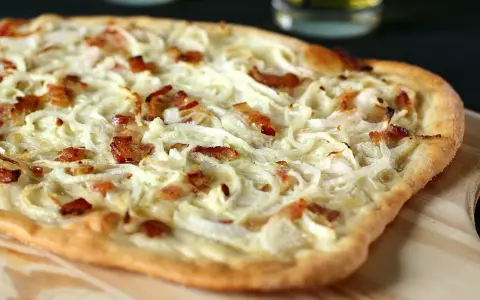 Flammekueche généreuse garnie d'oignons crus ou cuits et de lardons croustillants, prête à être dégustée.