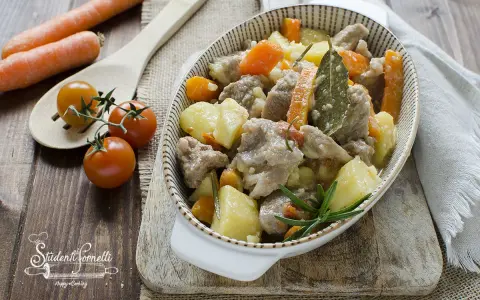 Un invitante spezzatino in bianco con patate e carote, guarnito con rosmarino e alloro. Accanto, pomodorini e carote fresche.