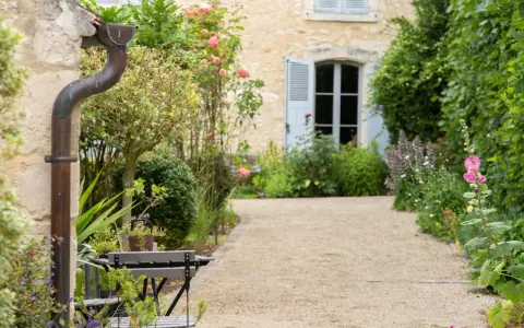 Un chemin de gravier serpente à travers un jardin anglais romantique, bordé de roses roses et de verdure luxuriante. Une maison en pierre avec des volets bleus se profile à l'arrière.