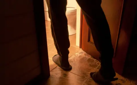 Nocne wstawanie do toalety, częste sikanie w nocy. Widok na nogi w kapciach i drzwi łazienki.
